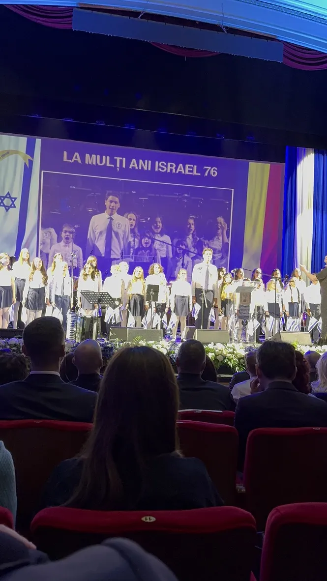 מקהלת ילדים בחולצות לבנות עם דגלי ישראל על במת האופרה, ומאחוריה מסך עם ברכה לישראל ברומנית